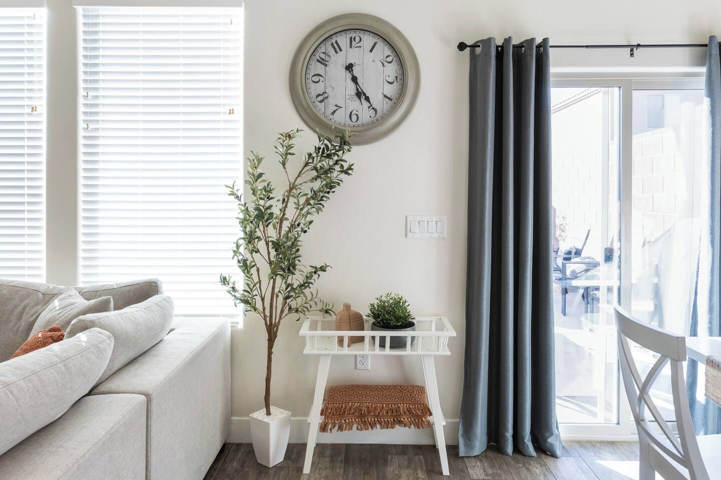 Ein Wohnzimmer mit einer Couch, einer Wanduhr, einem Blumentopf, einem weißen Regal mit Dekoration und Blick durch ein Fenster mit Vorhängen.