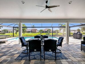 An outdoor patio with a table and chairs, a ceiling fan, and a view of a swimming pool and houses in the background. - Sous le Soleil- Canal, Heated Pool & Beach Gear! (Cape Coral)