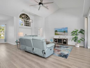 Ein geräumiges Wohnzimmer mit einem hellblauen Liegesofa, einem Deckenventilator, einem großen Fenster mit Blick und einem Bild an der Wand sowie einer Topfpflanze.