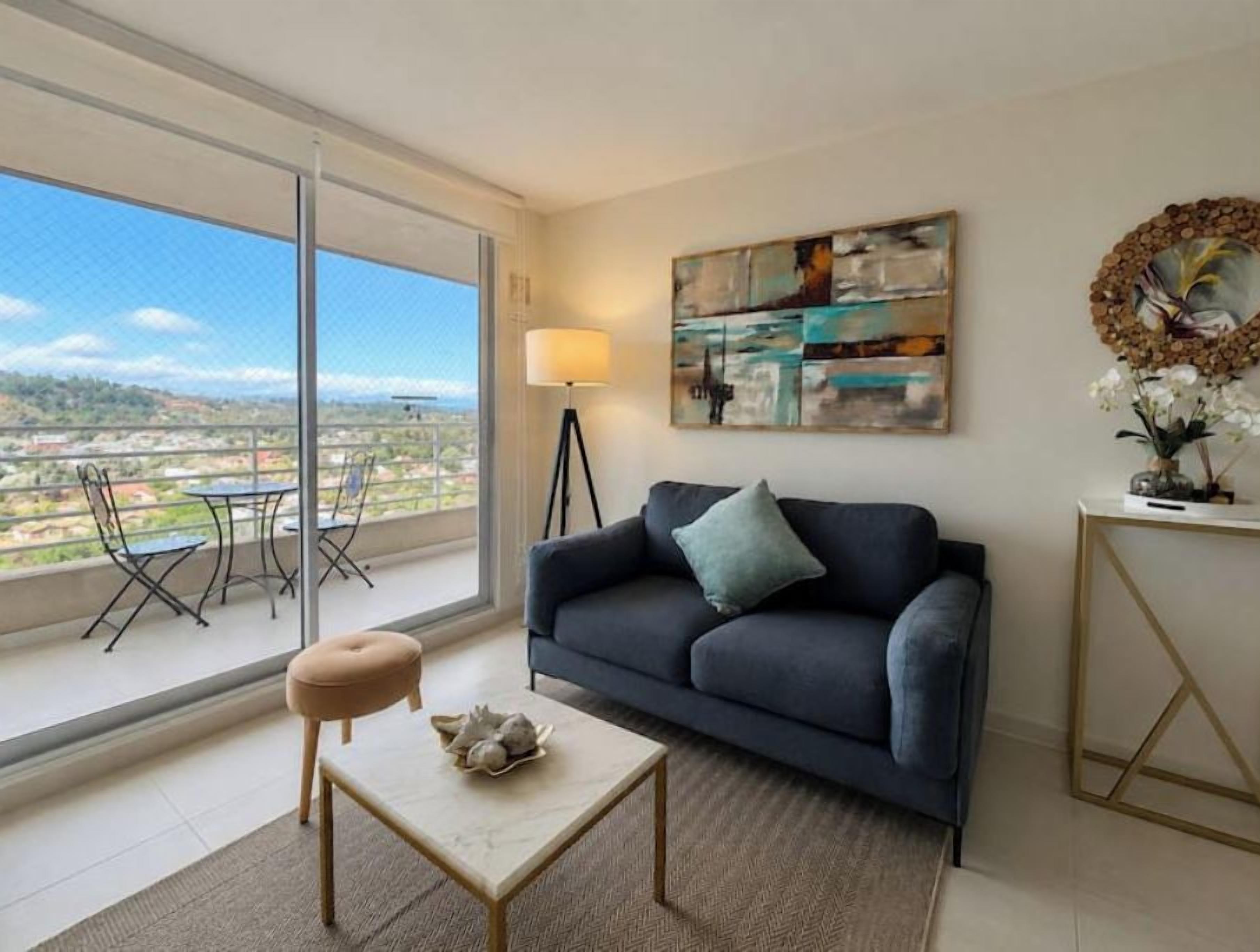 Una sala de estar moderna con un sofá, una mesita y una lámpara de pie. Se ve una ventana grande que ofrece vistas de la ciudad.