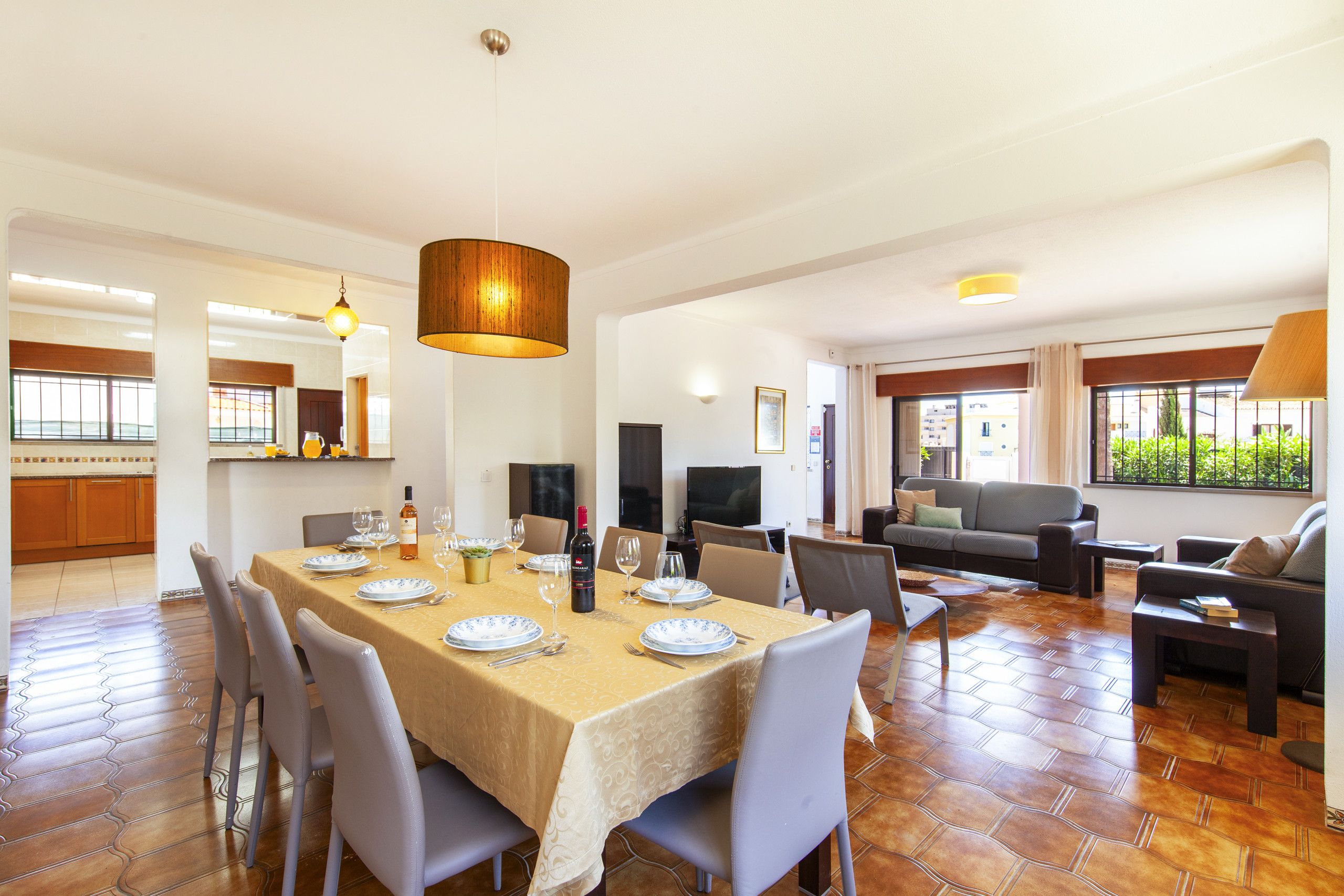 A spacious dining area with a large table set for six, surrounded by modern chairs. There is a television and a sofa in the living area, and large windows provide natural light.