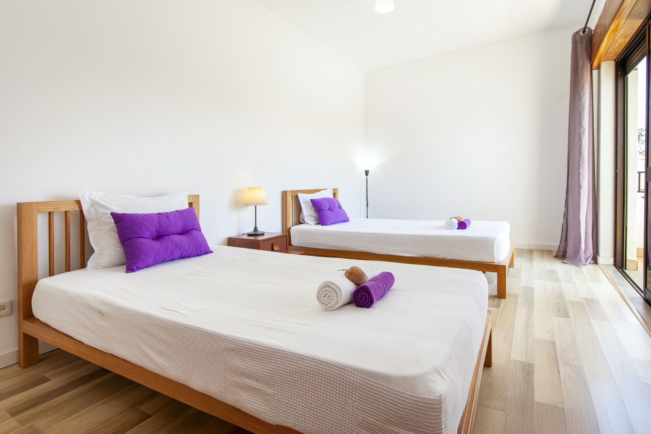 Habitación de hotel con dos camas, cada una con ropa blanca y almohadas moradas. Hay una lámpara en una mesita de noche y una ventana con cortinas.