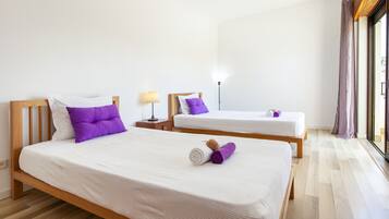 Habitación de hotel con dos camas, cada una con ropa blanca y almohadas moradas. Hay una lámpara en una mesita de noche y una ventana con cortinas.