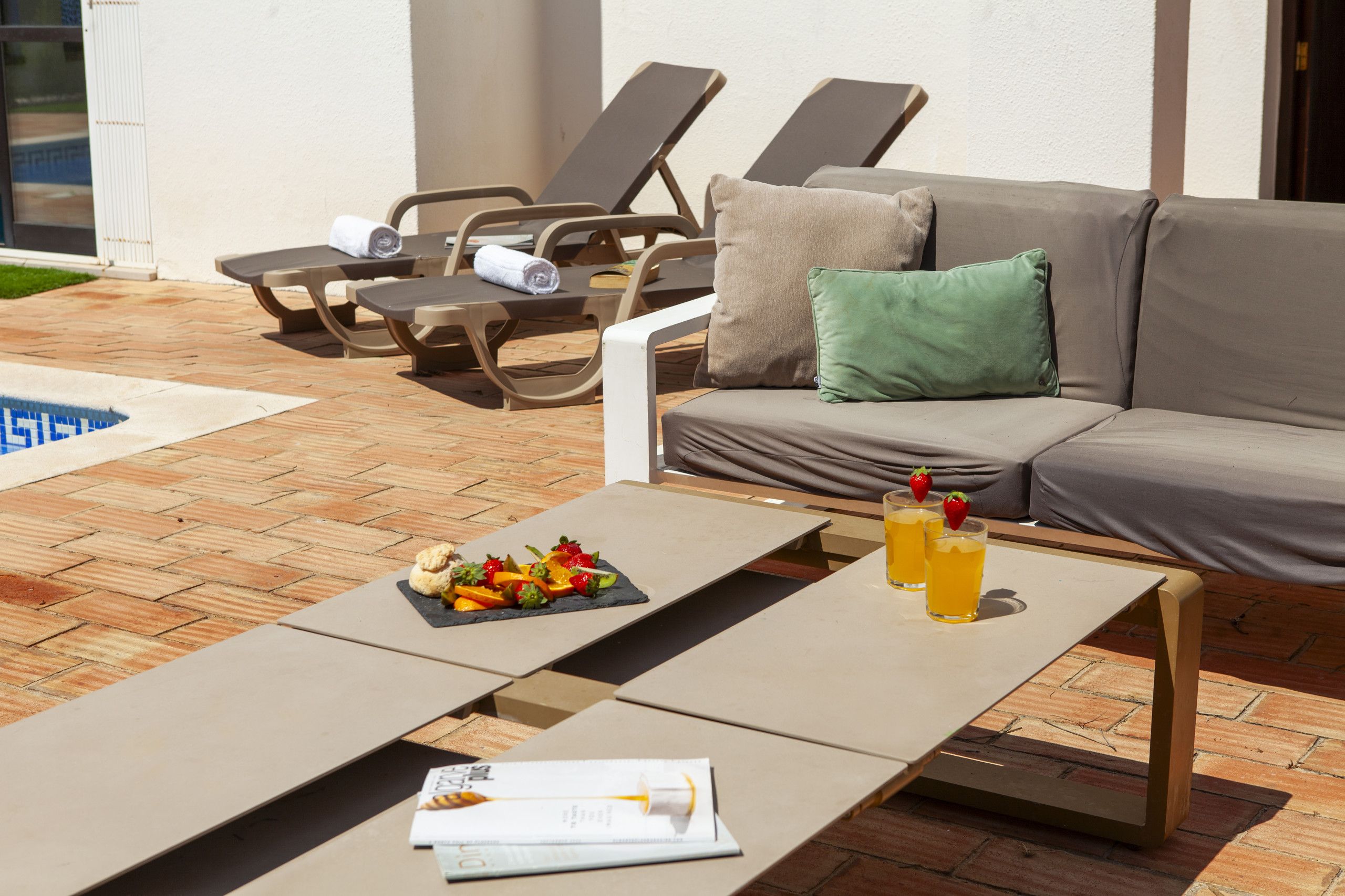 A poolside area with lounge chairs, a sofa, and a table with fruit and drinks.