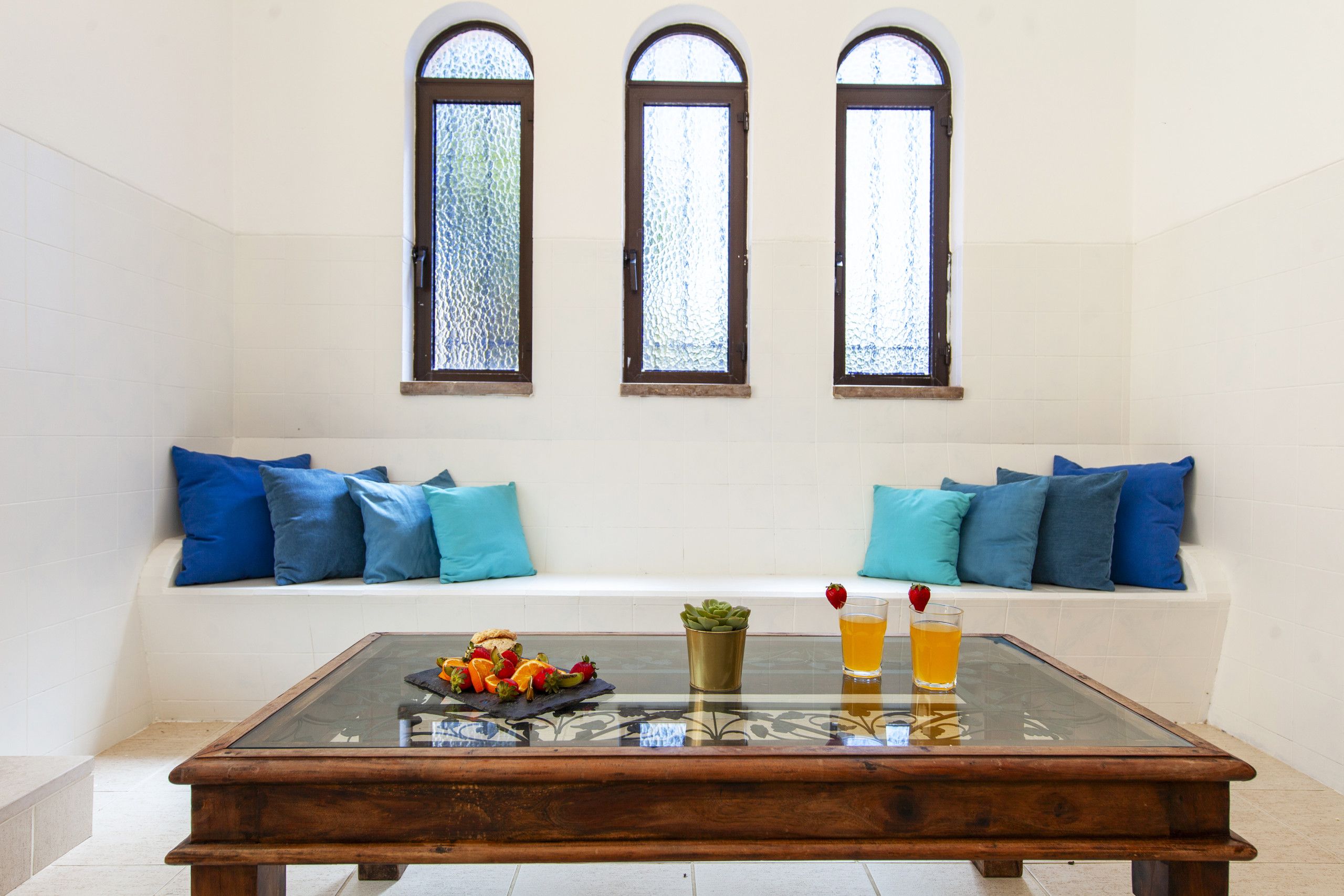 A room with a glass-top table, a bench with blue cushions, and three arched windows with frosted glass.