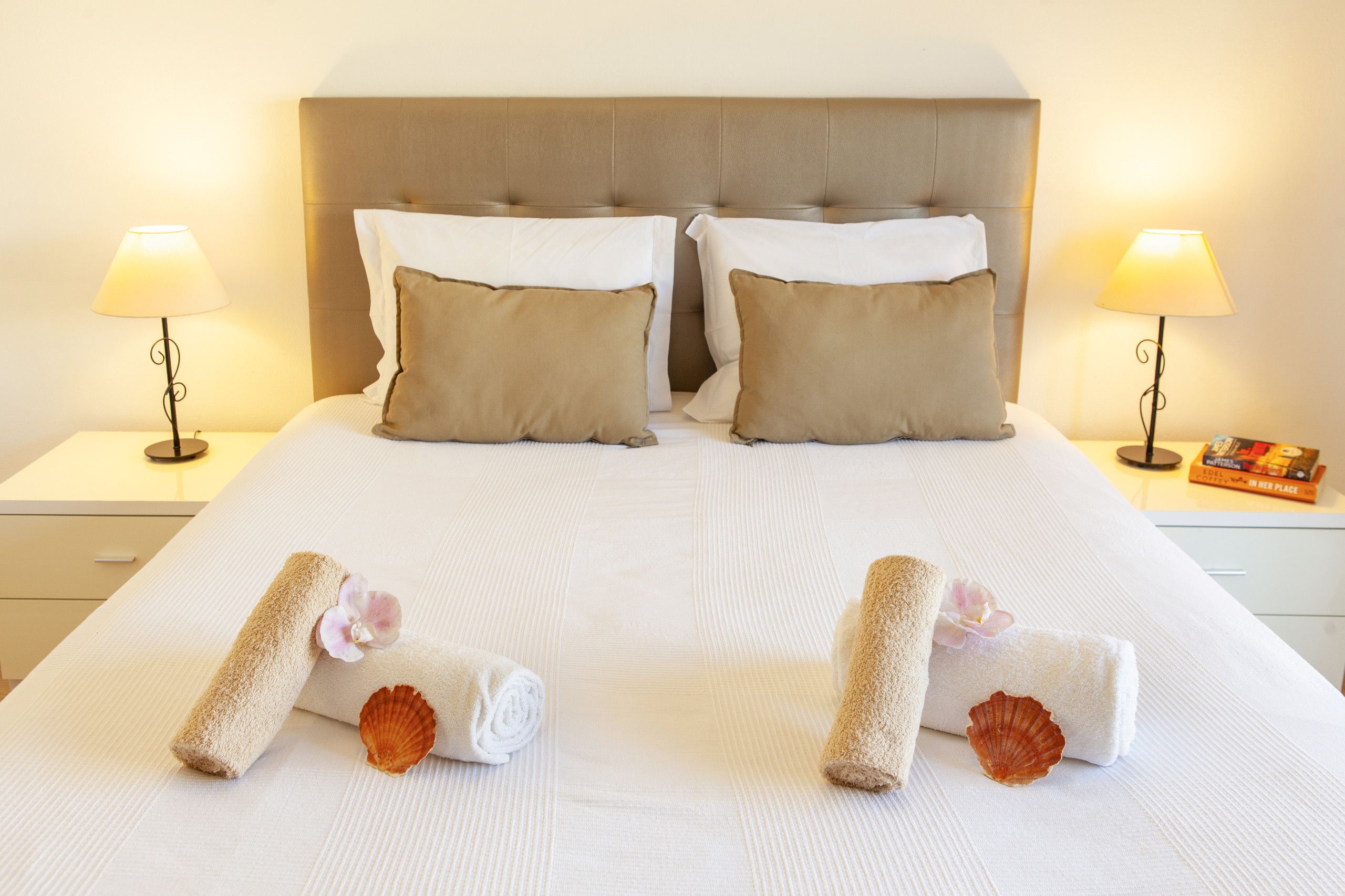 A neatly made bed with two pillows, two rolled towels, and a pair of seashells.