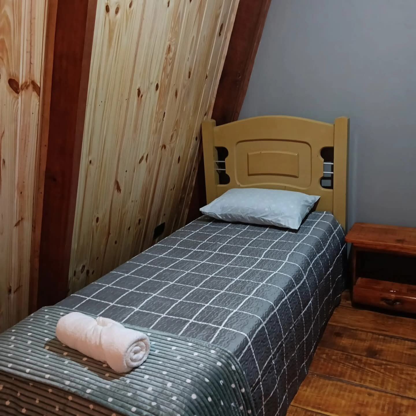 Quarto pequeno com uma cama de solteiro, cabeceira de madeira, colcha xadrez e toalha enrolada na cama.