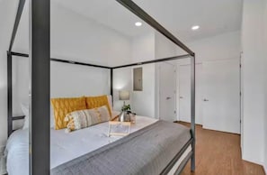 Ein Bett mit Baldachin, eine graue Tagesdecke, ein Nachttisch mit Lampe und eine kleine Pflanze.