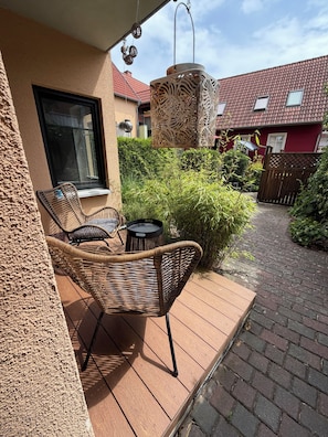 Une terrasse en bois avec des meubles en osier, un luminaire suspendu et, en arrière-plan, une vue sur un bâtiment au toit rouge.