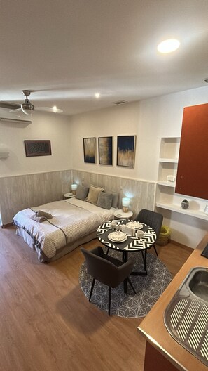 Ein kleines Hotelzimmer mit Bett, Esstisch mit Stühlen, einer Küchenzeile und einem Deckenventilator.
