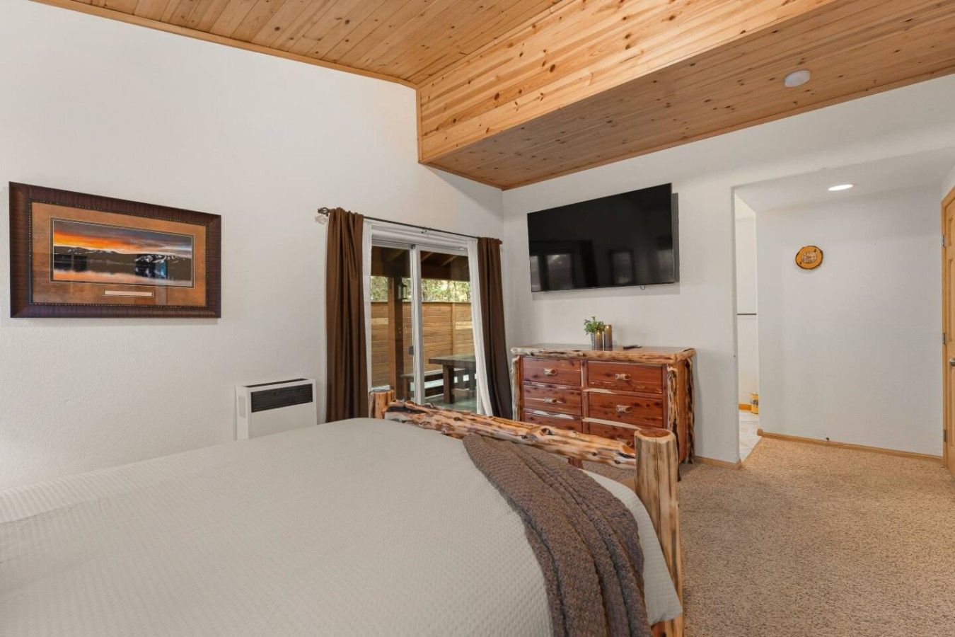 A bedroom with a bed, a wooden dresser, a television, and a sliding glass door leading to a balcony.