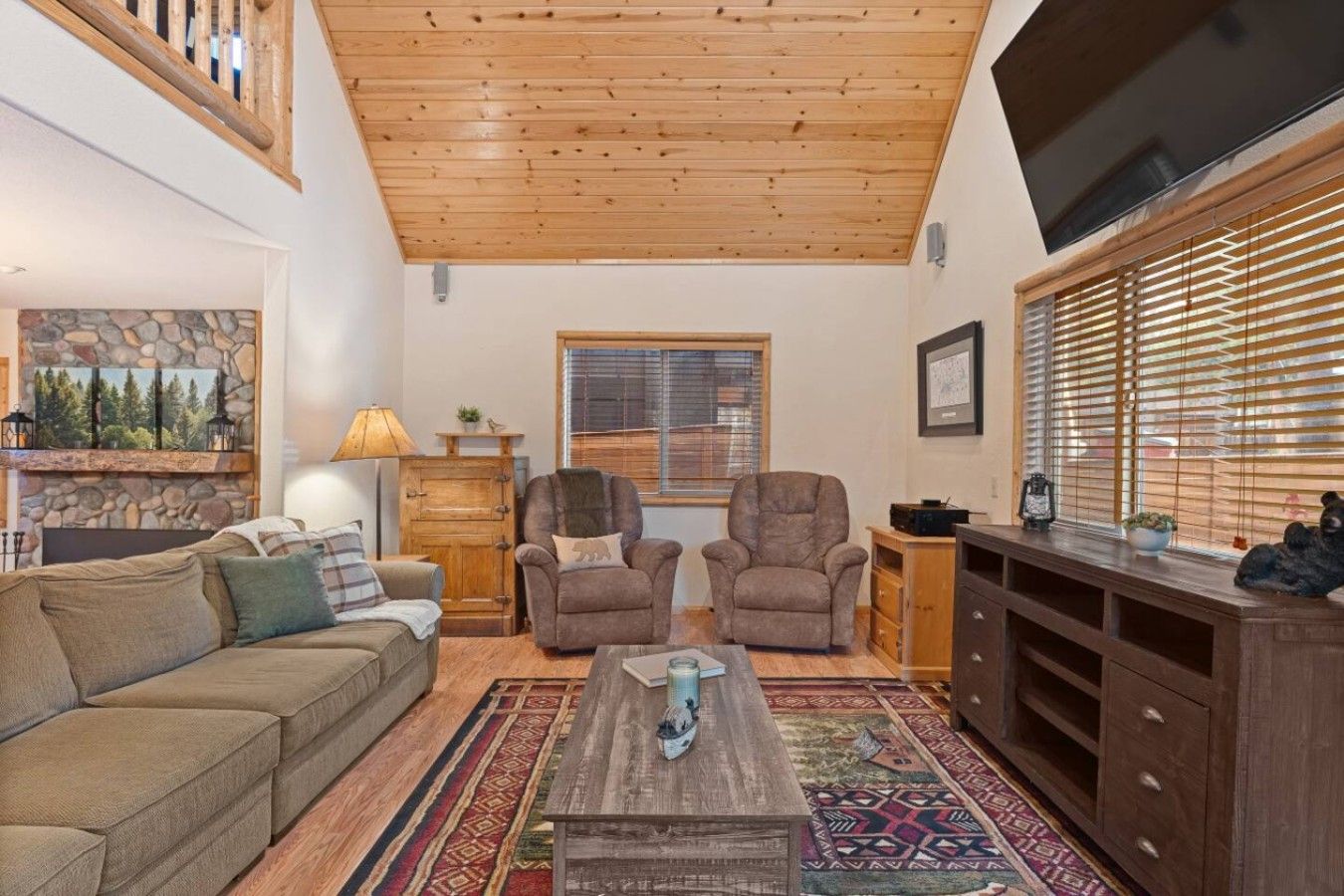 A living room with a wooden ceiling, a stone fireplace, a sofa, two armchairs, a coffee table, and a TV.