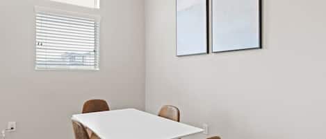 Una habitación con una mesa blanca y cuatro sillas, una ventana con persianas y dos cuadros en la pared.