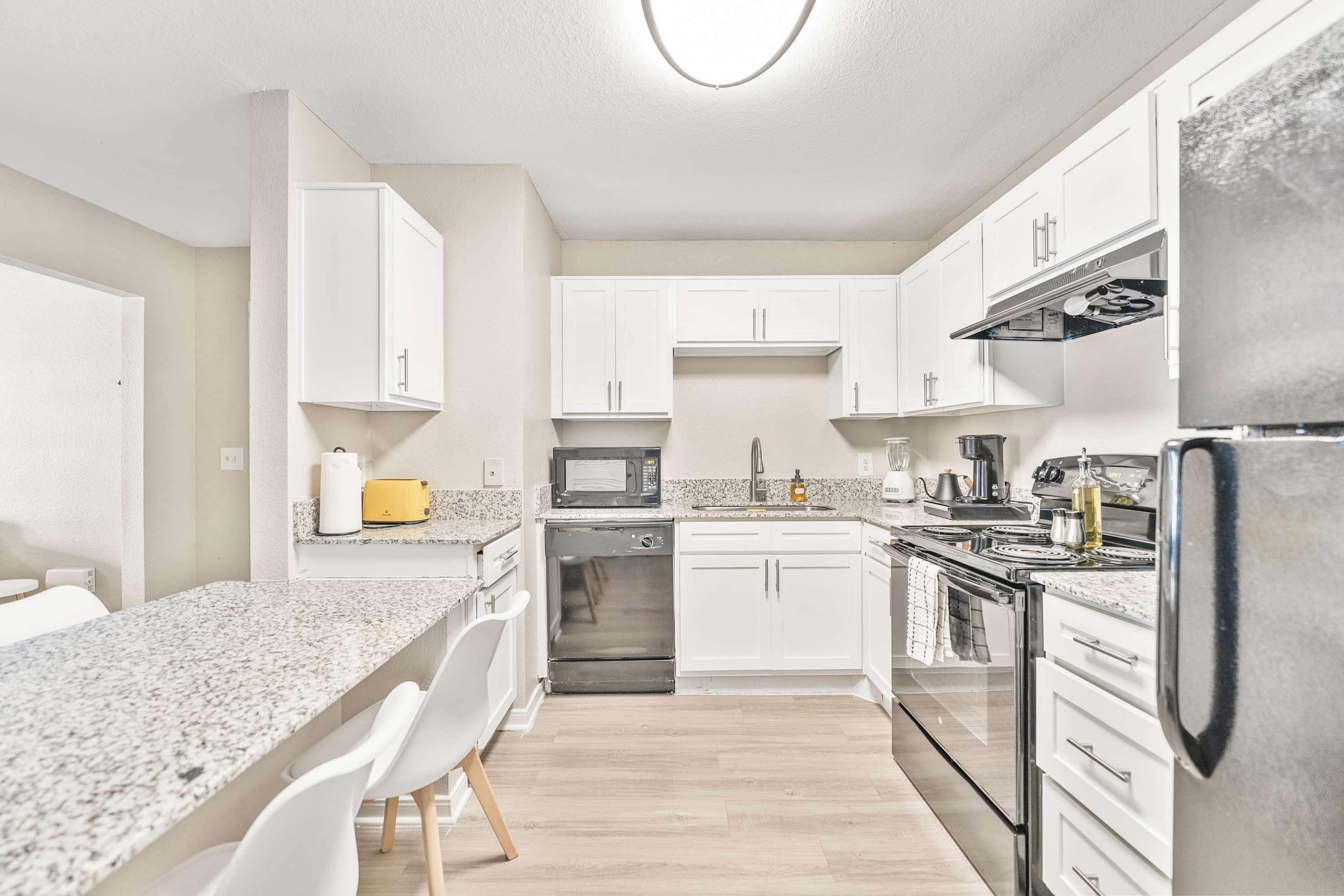 Een moderne keuken met witte kasten, een granieten aanrechtblad, roestvrijstalen apparatuur en een eetgedeelte met witte stoelen.