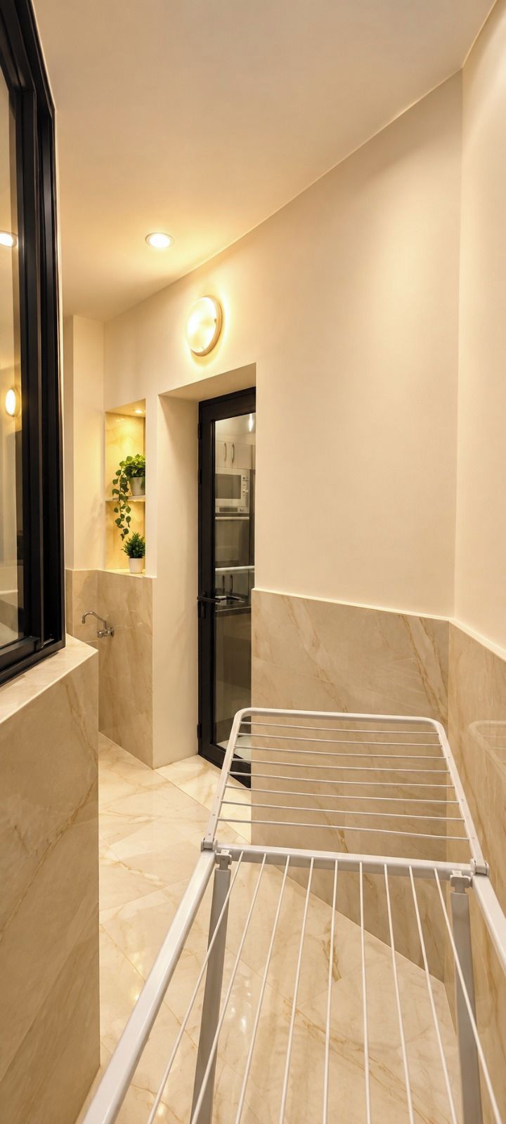 Un couloir étroit avec un étendoir à linge en métal, une porte en verre donnant sur une cuisine et un luminaire fixé au mur.