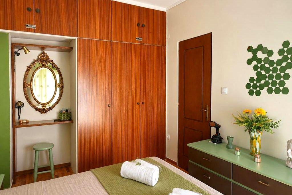 Une chambre à coucher avec un lit, un tabouret vert, une armoire en bois, un miroir et une commode ornée d’un vase de fleurs.