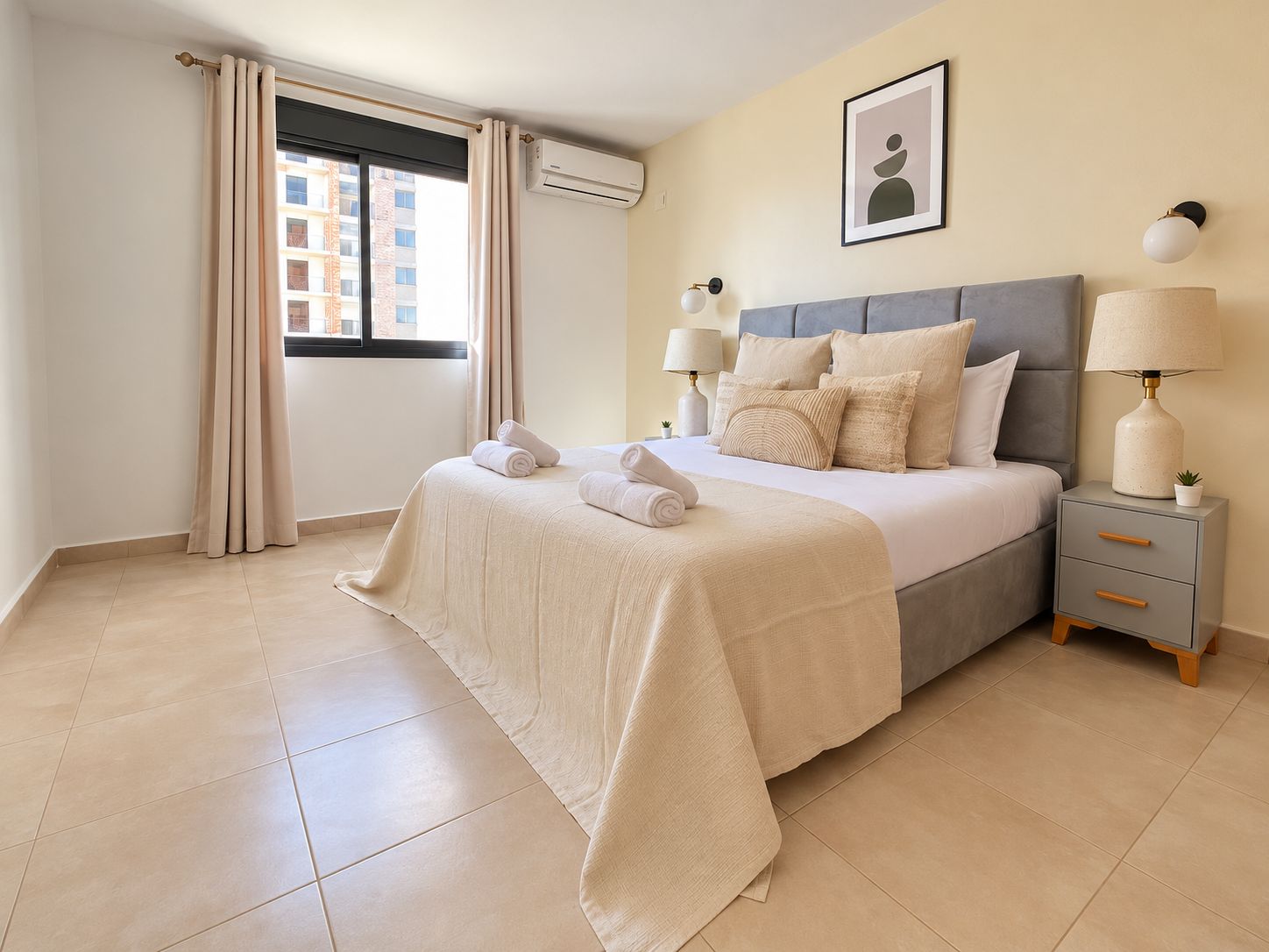 A hotel room with a bed, two bedside tables, a window with curtains, an air conditioning unit, and a framed picture on the wall.