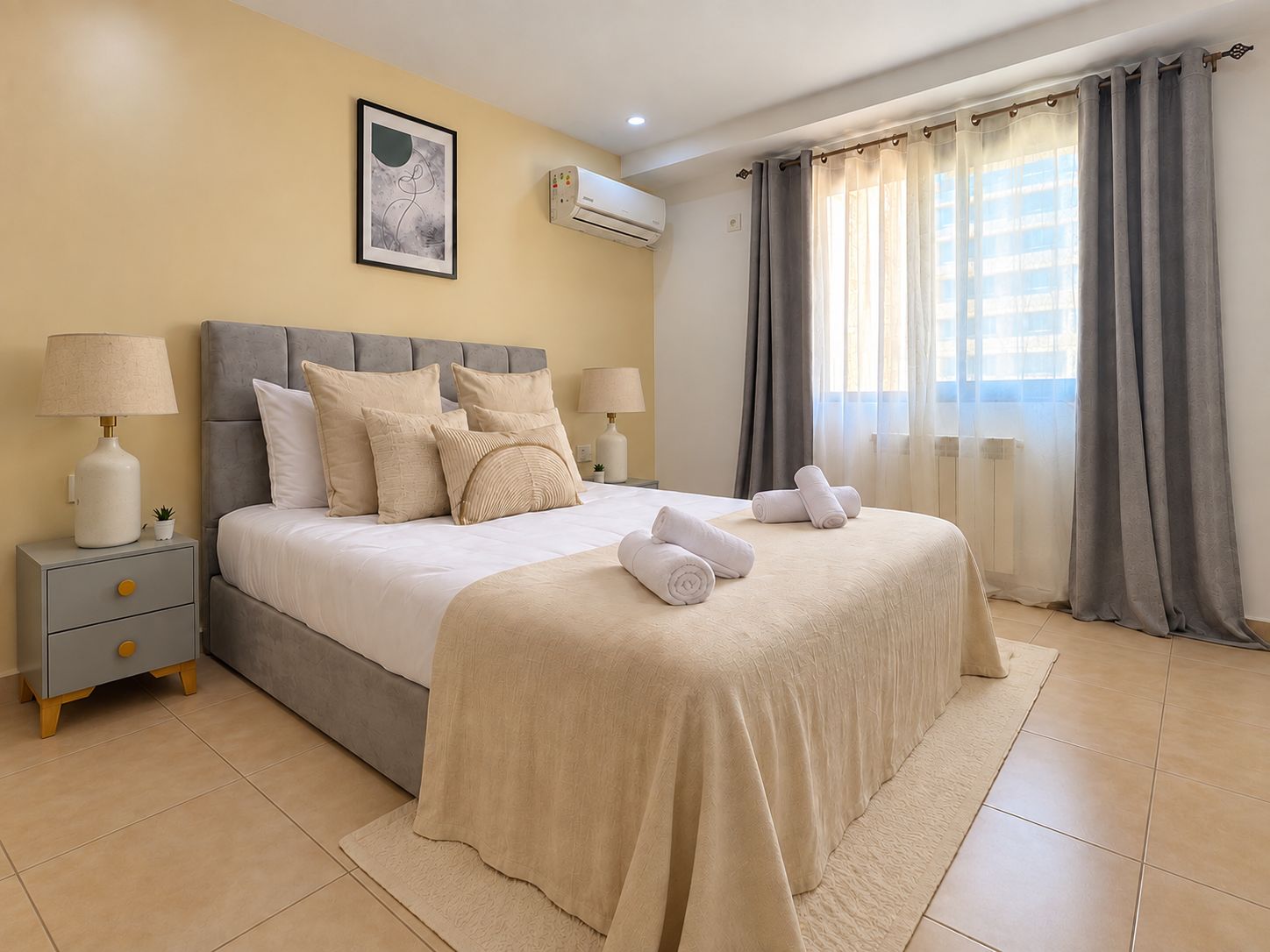 A hotel room with a bed, bedside tables, a wall-mounted air conditioner, and a window with curtains.