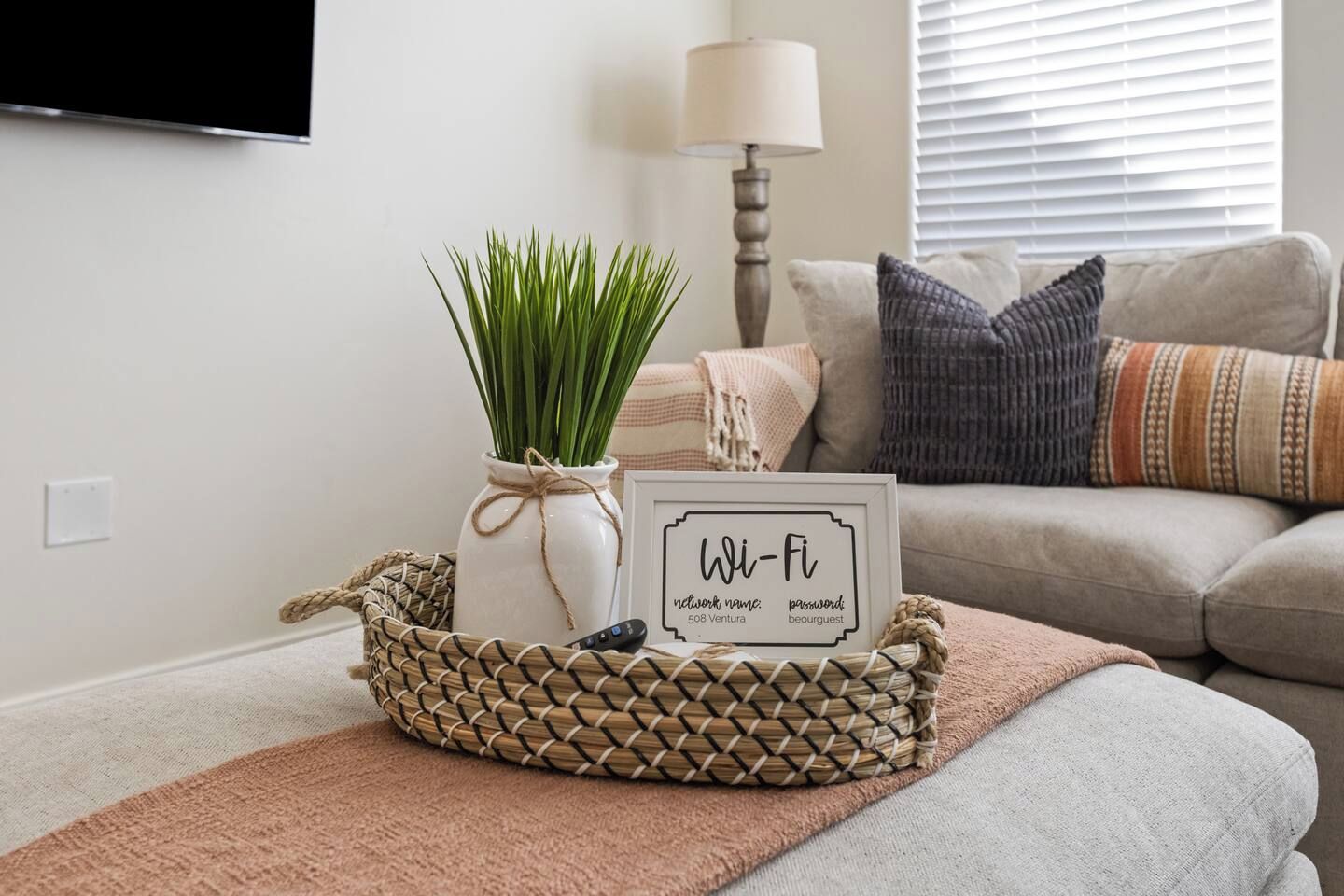 A living room with a TV mounted on the wall, a white vase with green plants, a framed Wi-Fi sign, and a woven basket with a remote control.
