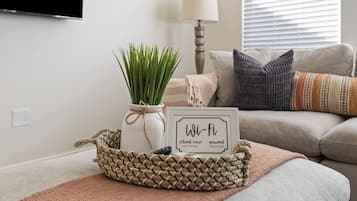 Un salon avec un téléviseur fixé au mur, un vase blanc contenant des plantes vertes, un panneau Wi-Fi encadré et un panier en osier contenant une télécommande.