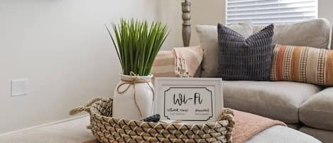 Un salon avec un téléviseur fixé au mur, un vase blanc contenant des plantes vertes, un panneau Wi-Fi encadré et un panier en osier contenant une télécommande.