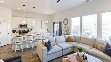 A modern living room with a sectional sofa, a dining area with white chairs, and a kitchen with white cabinets and stainless steel appliances.