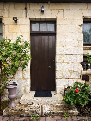 Un bâtiment en pierre avec une porte en bois sombre, une petite fenêtre ornée d’une grille décorative et un pot de fleurs contenant une fleur rouge.