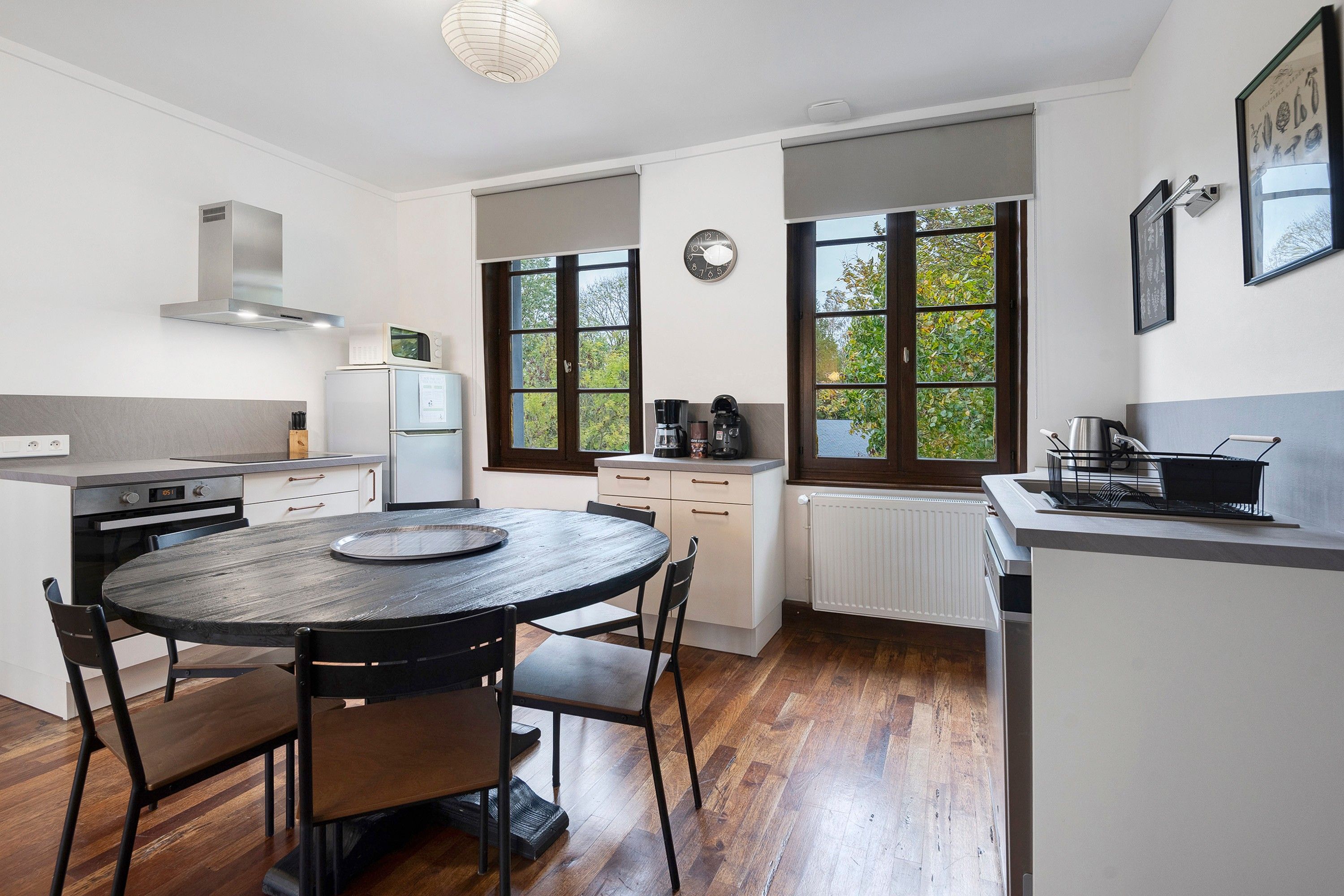 Een moderne keuken met een ronde eettafel, houten vloeren en grote ramen met uitzicht op groen.