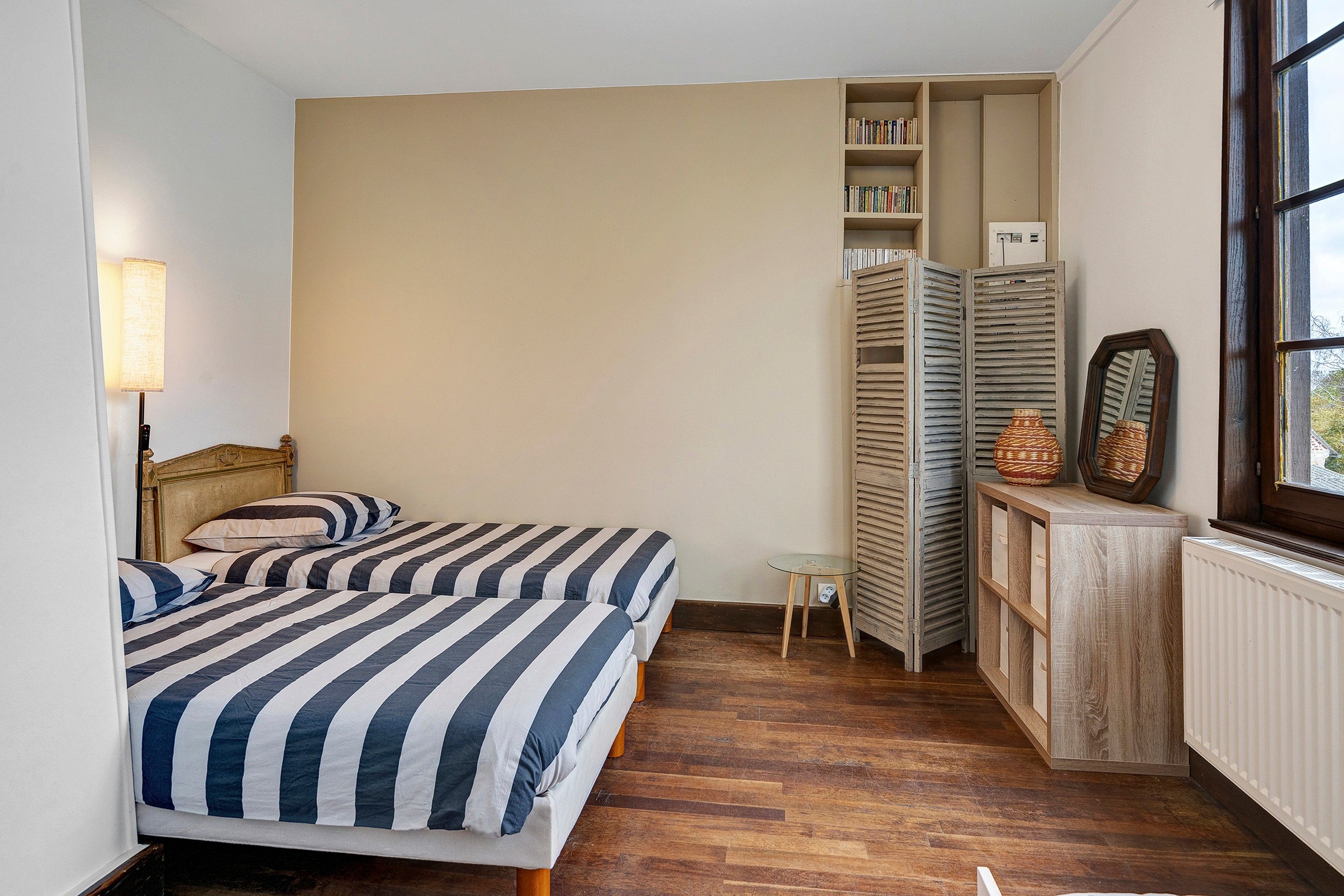 Un dormitorio con dos camas, una mesita de noche, una lámpara, una estantería y una ventana.