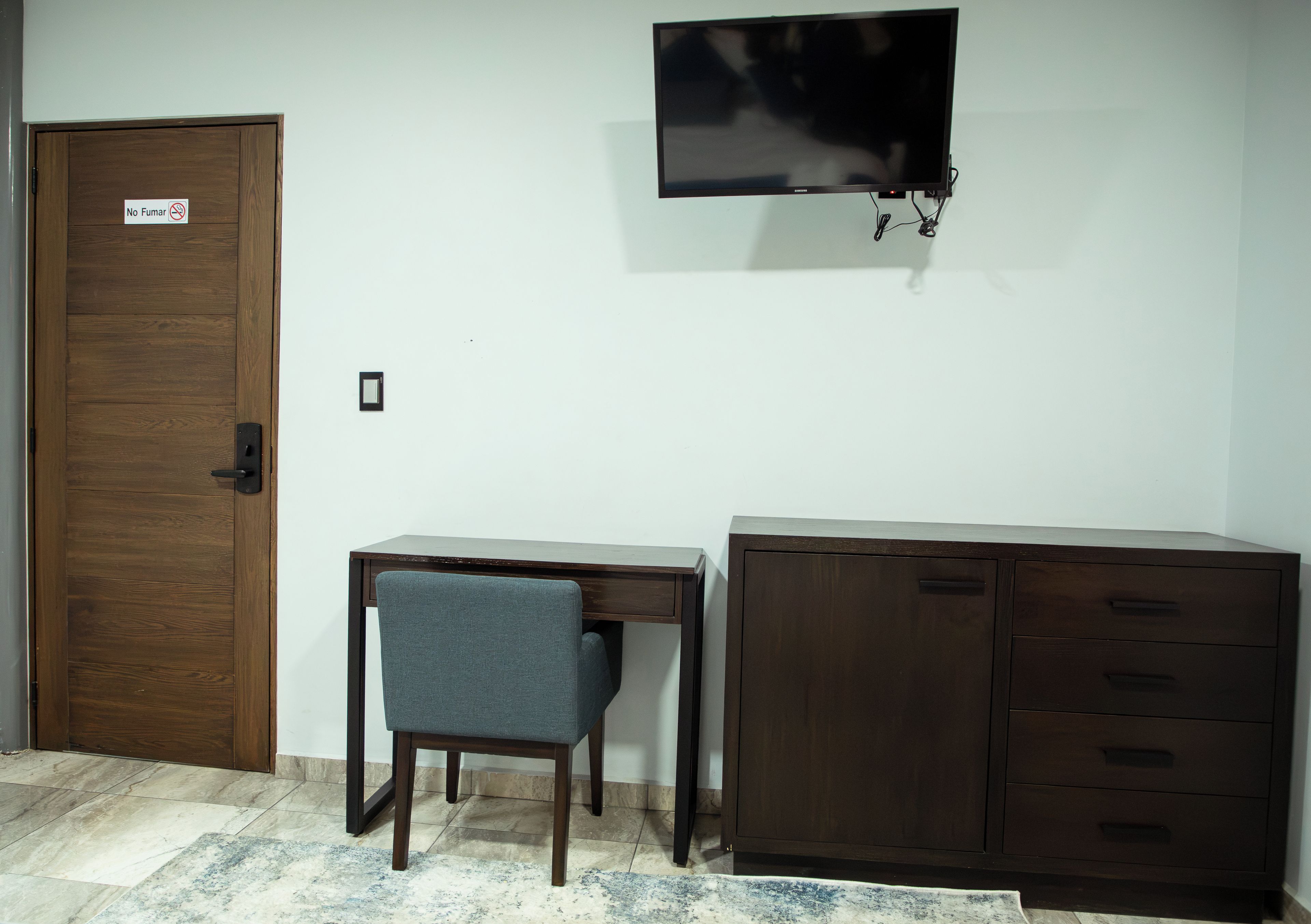 Una habitación con puerta de madera, un escritorio con silla, un televisor montado en la pared y un archivador.