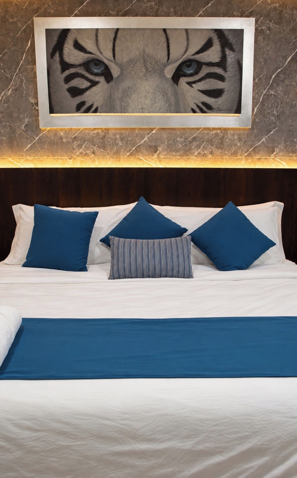 Una cama con ropa de cama y almohadas azules, un cuadro con imagen de un tigre en la pared, y una pared con textura similar al mármol.