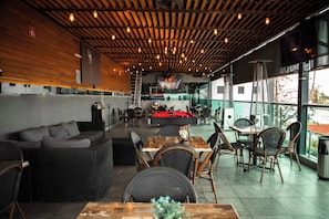 An indoor dining area with wooden ceiling beams, tables, chairs, and a pool table.