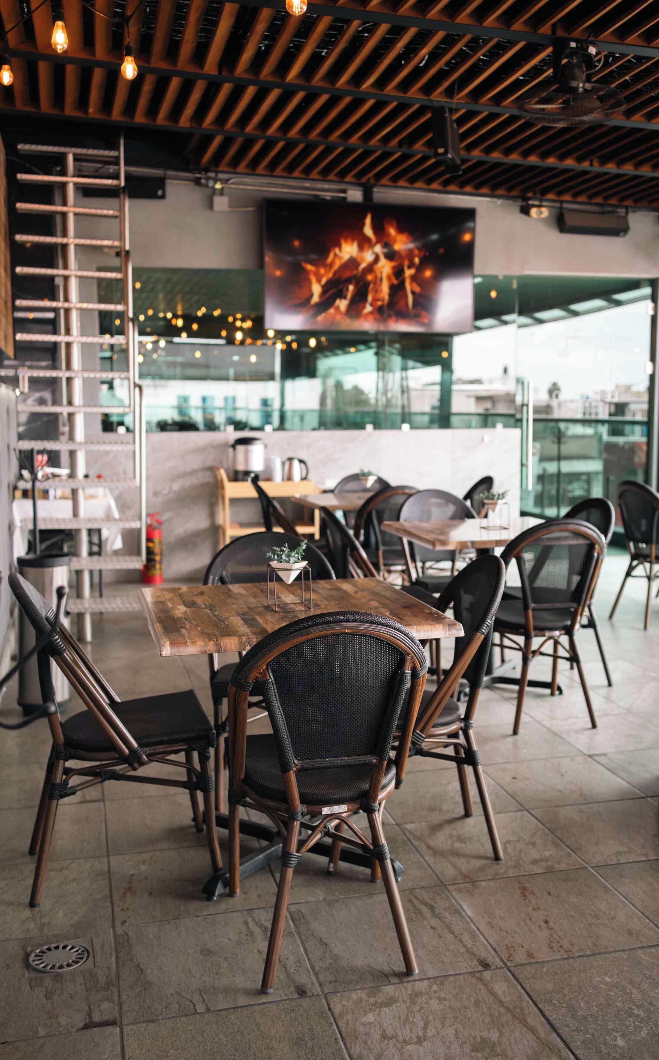 An outdoor dining area with wooden tables and wicker chairs, a fire feature on the wall, and a view of the cityscape.