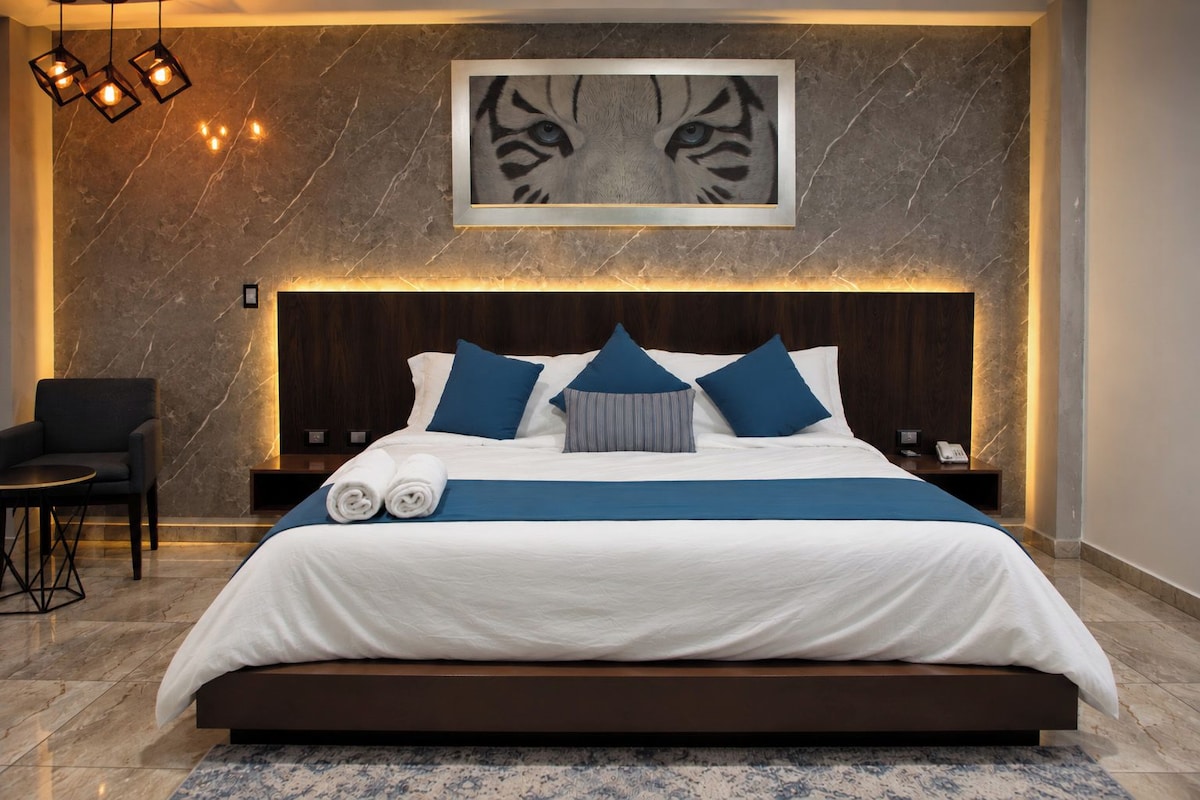 A modern hotel room with a large bed, a headboard, a framed artwork, a chair, and a small table.