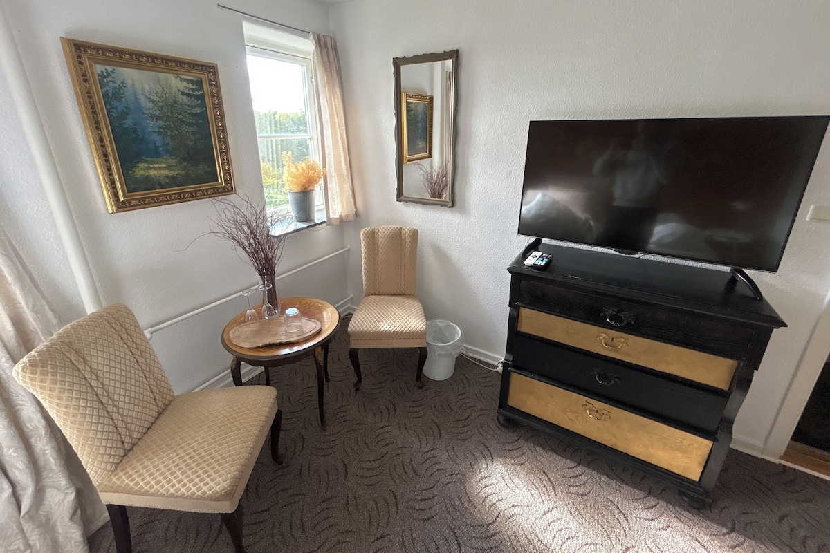 A hotel room with a flat-screen TV, a sofa, a small round table with a vase, and a window with curtains.
