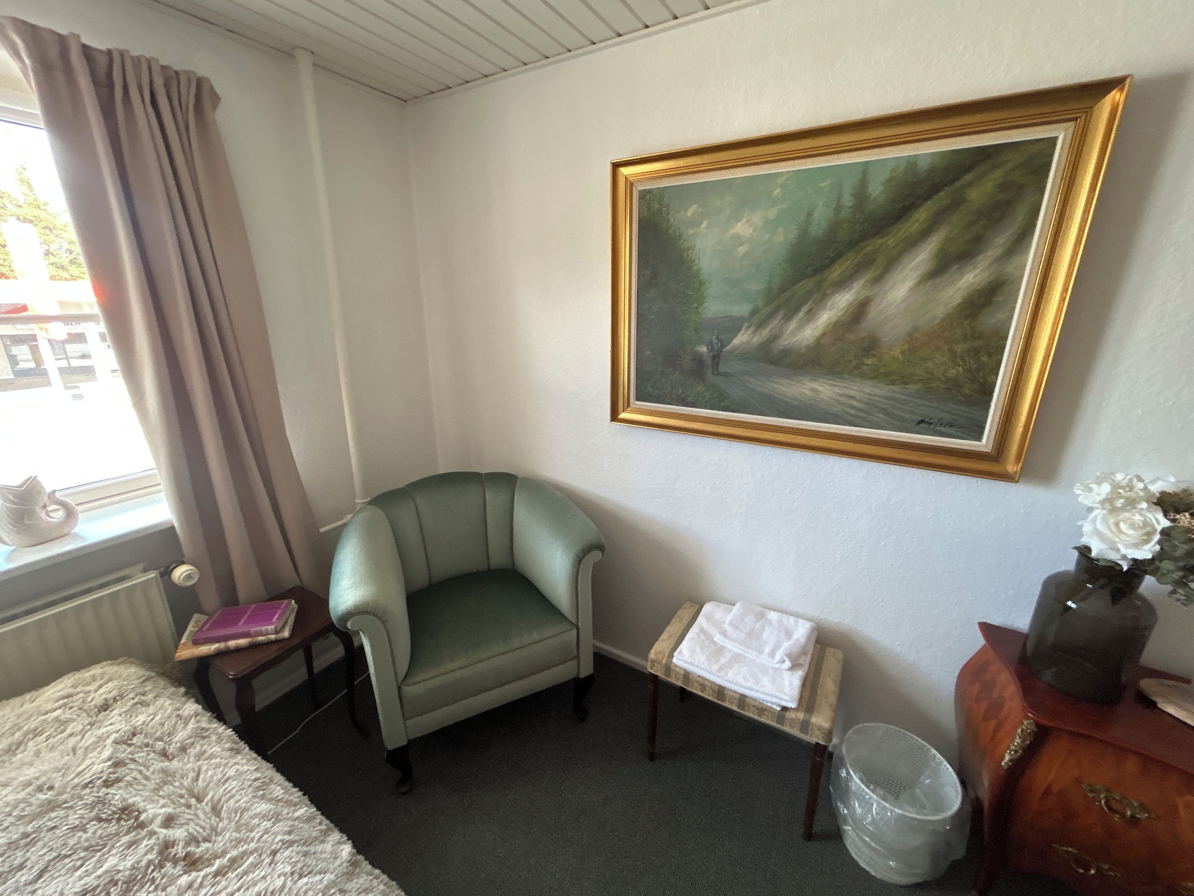 Et værelse med en grøn lænestol, et billede på væggen, et vindue med gardiner, et lille bord med en bog og en vase med blomster.