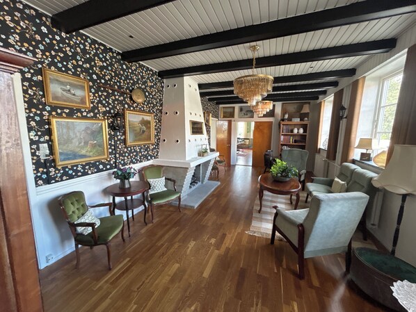 A spacious living room with wooden flooring, a fireplace, and a chandelier. There are several chairs and a sofa arranged around the room.