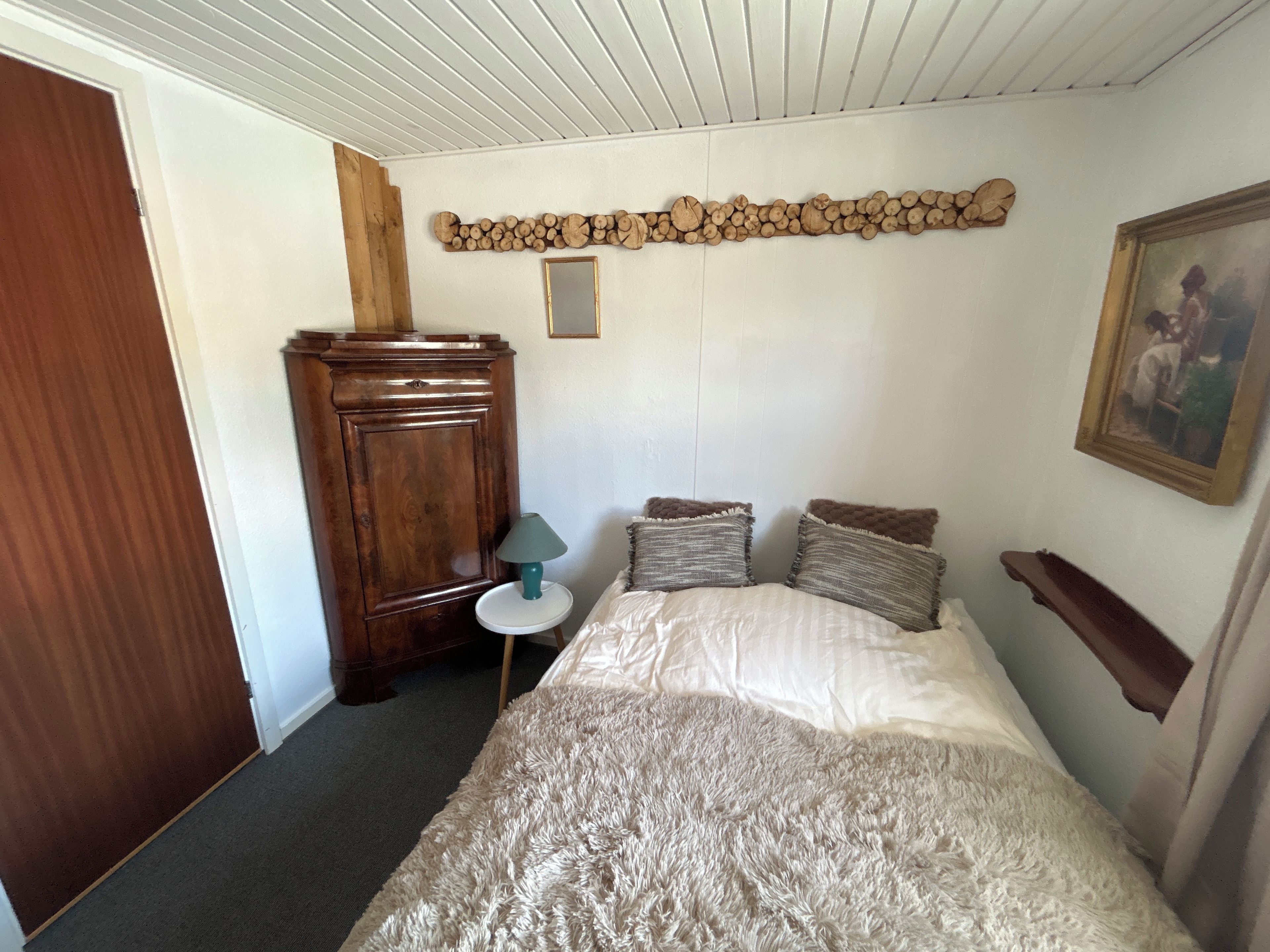 A bedroom with a bed, a wooden wardrobe, a small table with a lamp, and a painting on the wall.