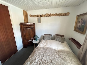 A bedroom with a bed, a wooden wardrobe, a small table with a lamp, and a painting on the wall.