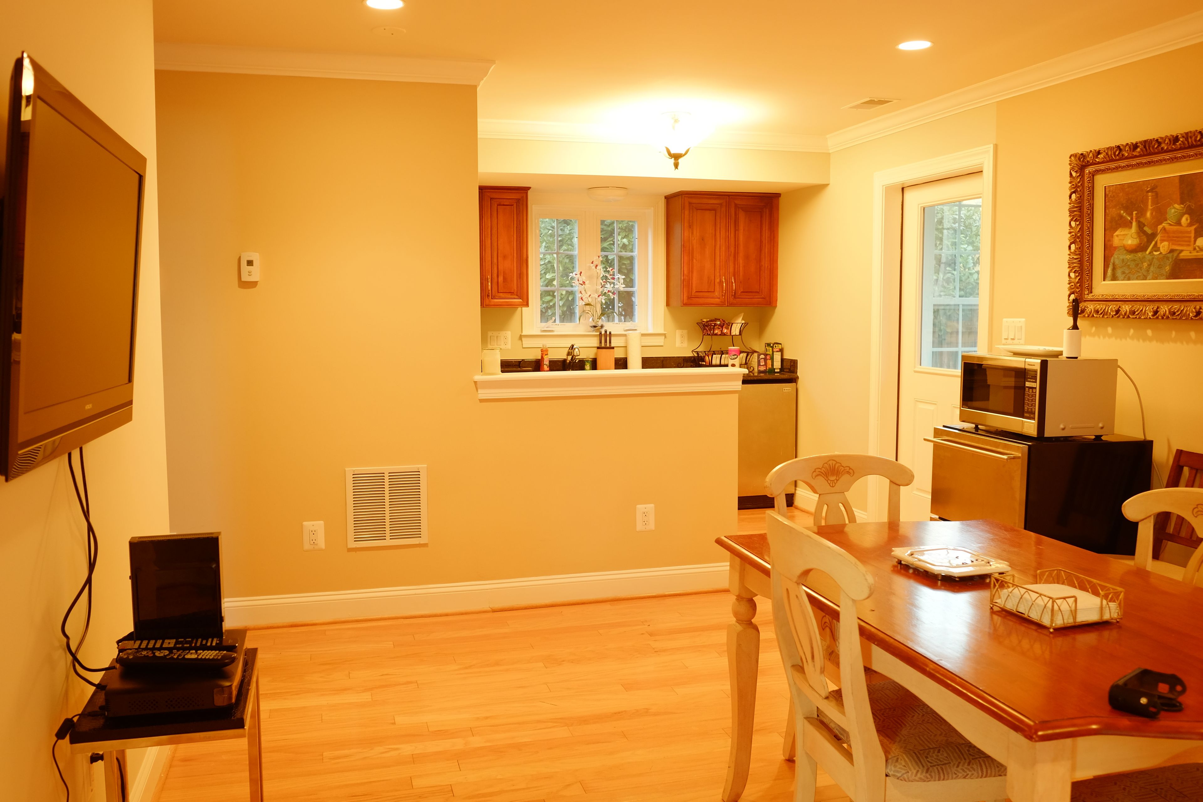 A kitchen with wooden cabinets, a dining table with chairs, a television mounted on the wall, and a microwave.