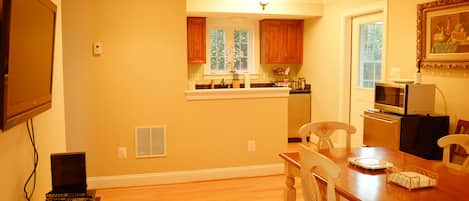 A kitchen with wooden cabinets, a dining table with chairs, a television mounted on the wall, and a microwave.