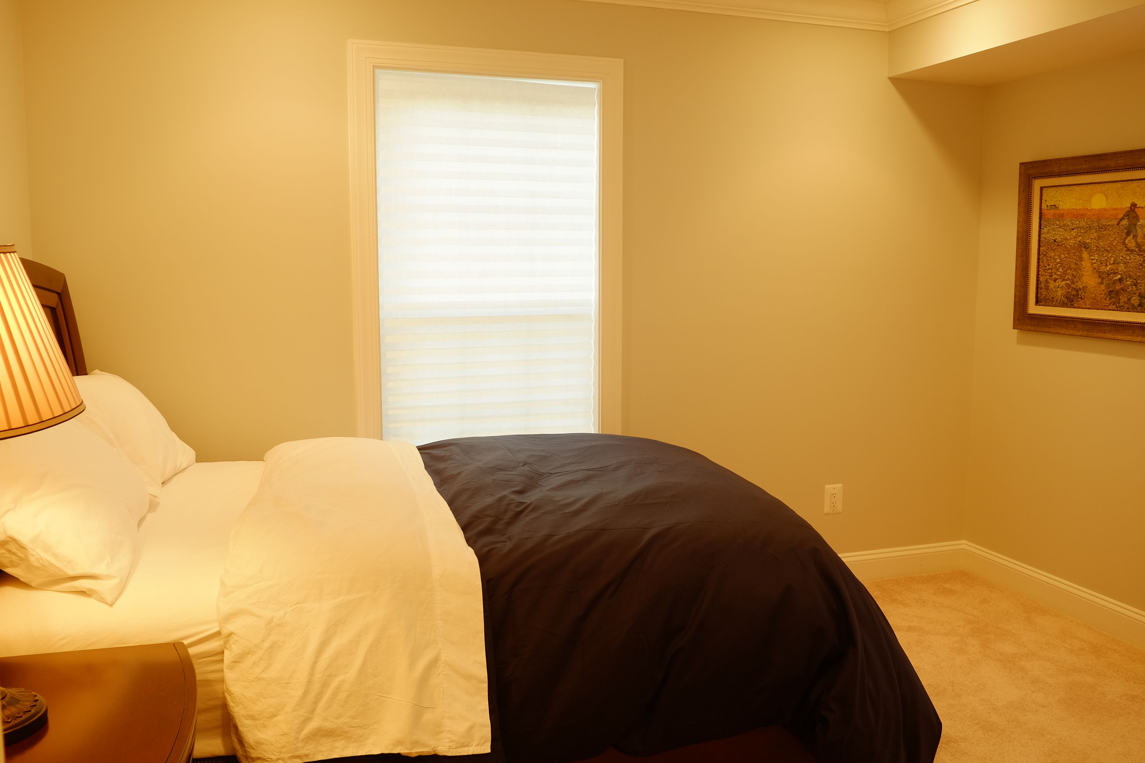 Um quarto com cama, abajur, janela com persianas e um quadro na parede.