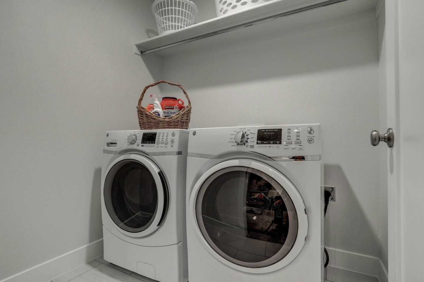 Una sala de bugaderia amb dues rentadores de càrrega frontal i un cistell a una estant a sobre.