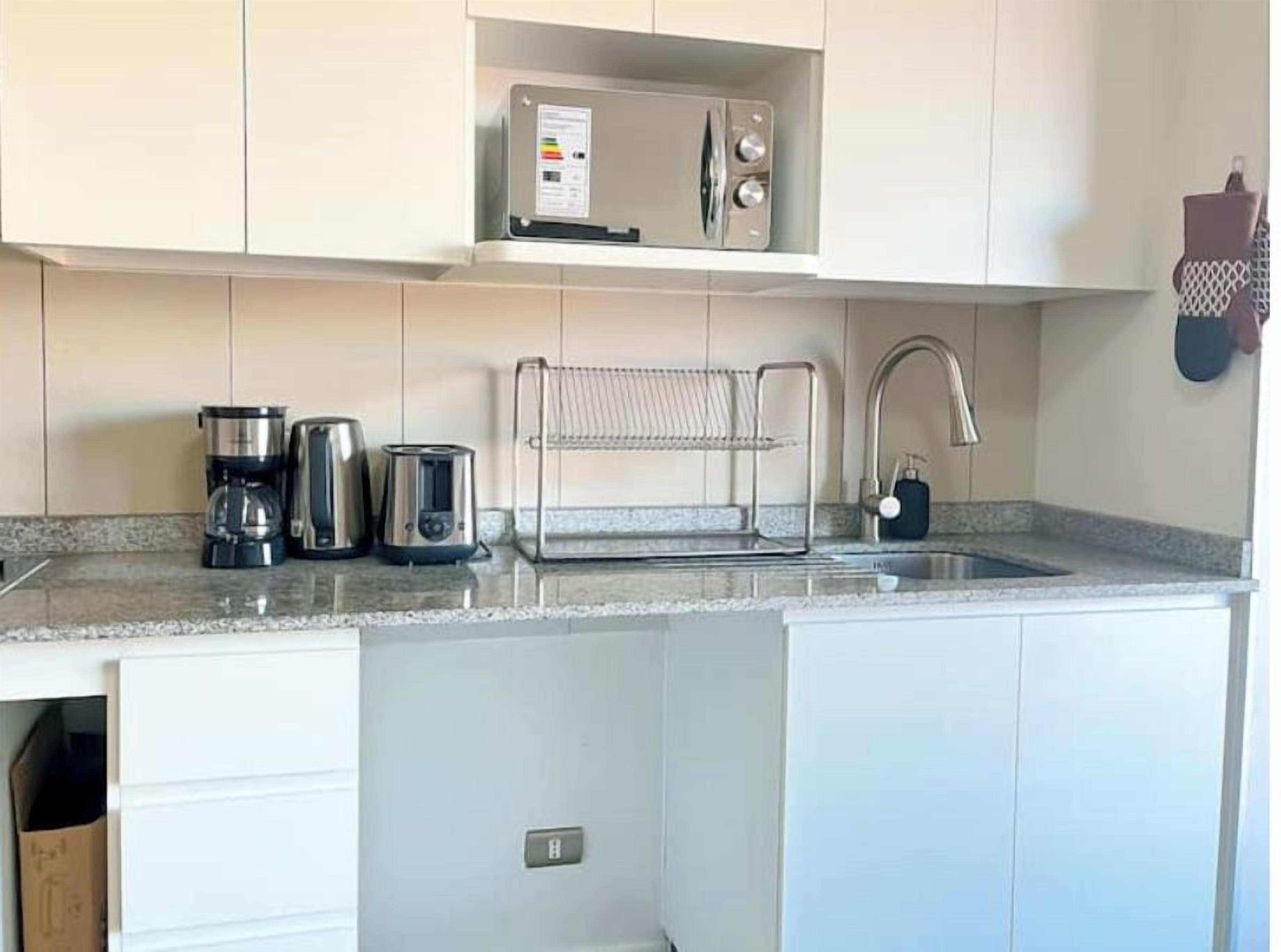 Cocina compacta con microondas, cafetera y hervidor sobre la encimera, un perchero y un lavabo con grifo.
