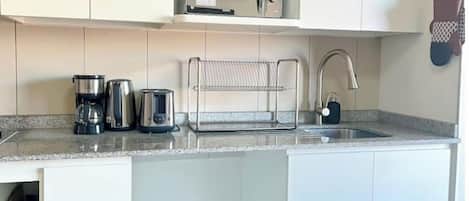 A compact kitchen with a microwave, coffee maker, and kettle on the countertop, a hanging rack, and a sink with a faucet.