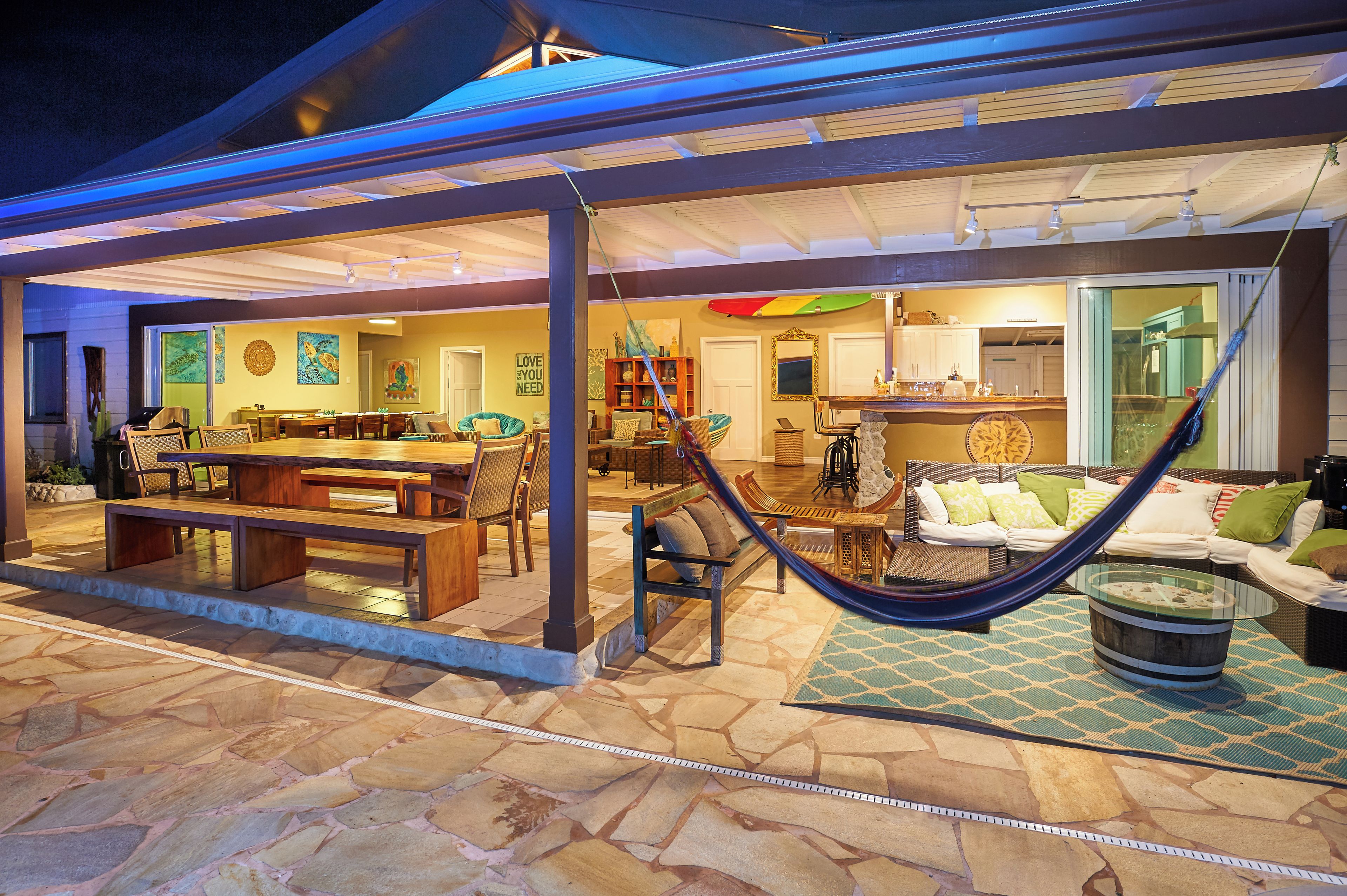 A covered outdoor area with a dining table, chairs, a hammock, and a kitchen area.