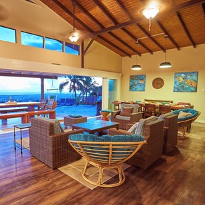 A spacious lounge area with wicker furniture, a view of the ocean, and a high ceiling with wooden beams.