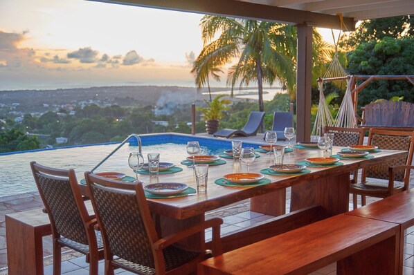 A dining table set for six with chairs, overlooking a pool and a scenic view of a coastal landscape.