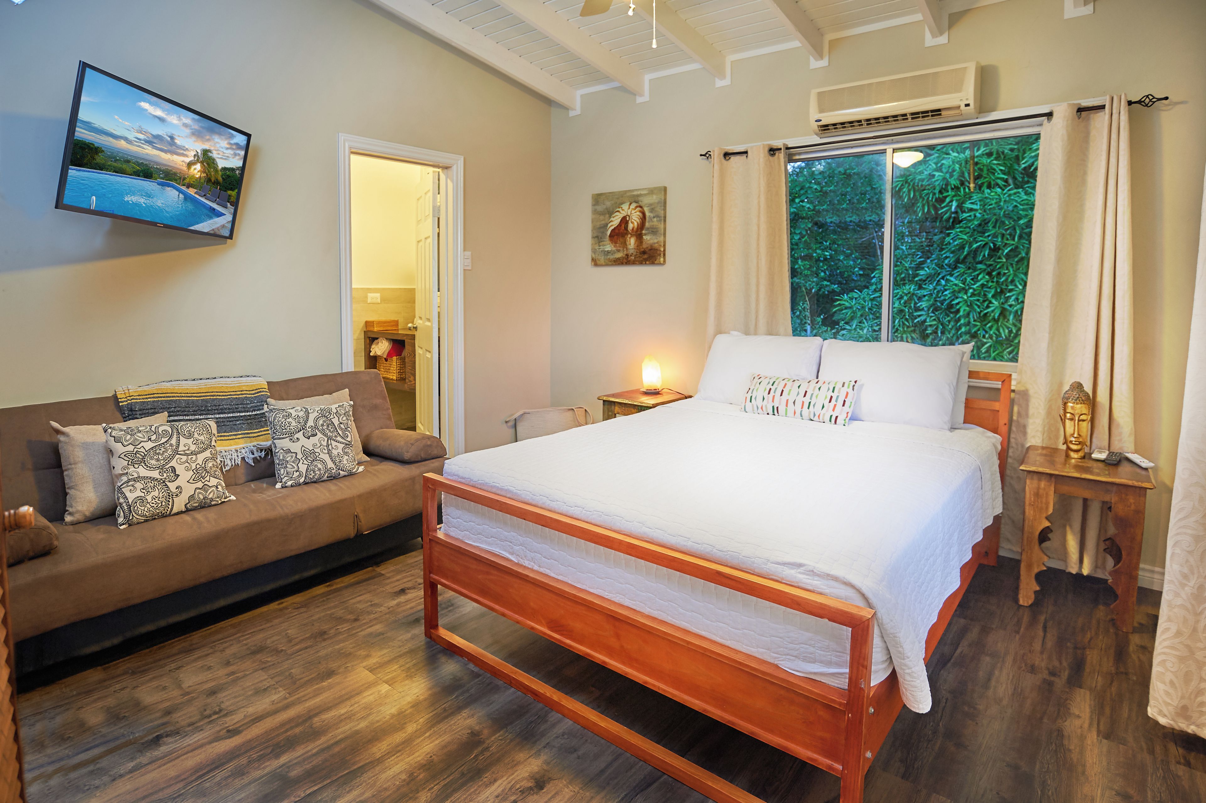 A bedroom with a bed, a sofa, a bedside table, a lamp, a painting, and a window with curtains.