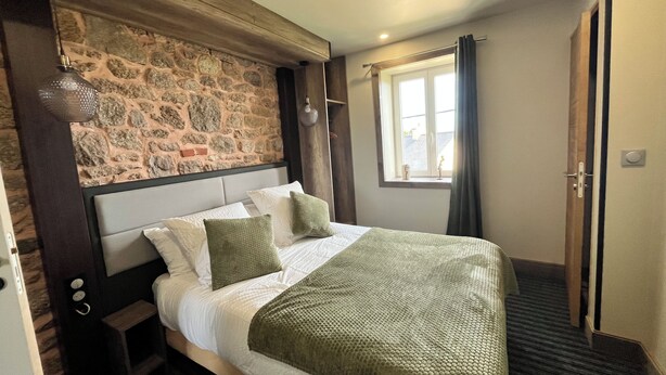 A bedroom with a stone wall, a bed with white bedding and green pillows, a window with curtains, and a door. - Vrbo Property (Vicq-sur-Mer)